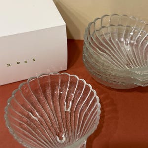 May include: A set of clear glass bowls shaped like seashells with ribbed textures and three small feet. A white box with the text "host" printed in small green letters is positioned behind the stacked and individual glass bowls.