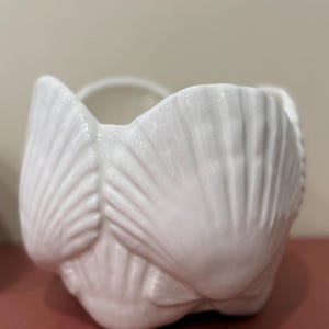 May include: A white ceramic bowl featuring a molded shell design with a glossy finish. The exterior surface is composed of multiple overlapping scallop shell shapes, creating a textured, scalloped rim and a rounded, bulbous base structure.