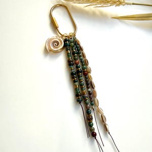 May include: A gold-colored keychain with a seashell charm and beaded tassels. The tassels feature green, brown, and tan beads, accented with gold-colored spacers. The seashell charm is white and tan.
