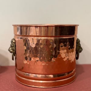 May include: A cylindrical copper bucket featuring a hammered texture on the exterior surface. The vessel includes two decorative metal handles shaped like lion heads, positioned on opposite sides of the polished metallic container.