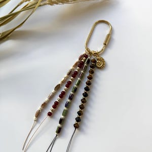May include: A gold-colored keychain with four beaded strands in various colors. The beads include cream, red, green, and brown tones. A small gold charm in the shape of a spiral shell hangs from the keychain.