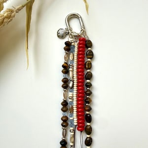 May include: A beaded keychain with a silver clasp and charm. The keychain features strands of brown, red, white, and blue beads. The beads are various shapes and sizes, including round, oval, and cylindrical. The keychain is a decorative accessory.