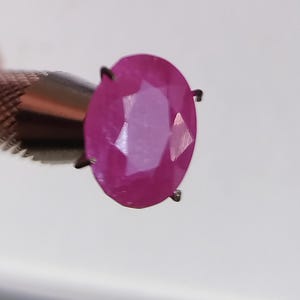 Könnte beinhalten: Ein facettierter, oval geschliffener Rubin-Edelstein, der von einem Metallwerkzeug gehalten wird. Der Rubin hat einen leuchtenden rosa-roten Farbton mit sichtbaren inneren Facetten. Das Metallwerkzeug hat einen strukturierten Griff und kleine Zinken zur Sicherung des Edelsteins.