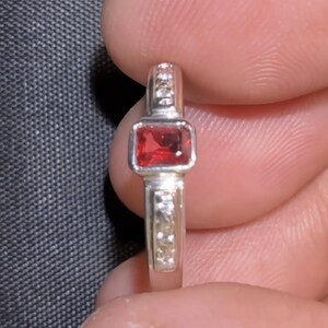 May include: A silver ring with a square, red gemstone at the center. The band is accented with small, clear stones. The ring is held between fingers against a dark background.