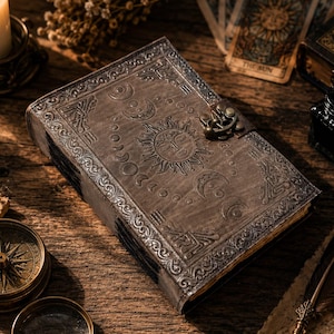 May include: A rectangular journal with a dark brown cover featuring embossed sun and moon phase designs. The book is secured with a metallic latch and has intricate border patterns. The surface of the cover appears textured and aged in appearance.