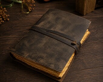 May include: A dark brown leather journal with a weathered, matte texture and a wrap-around leather cord closure. The journal features thick, deckled-edge pages with a light tan, aged appearance, visible along the side of the closed book.