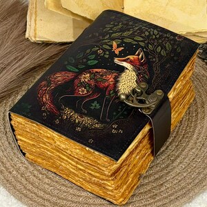 May include: A leather-bound journal with a fox illustration. The cover features a detailed image of a fox with floral patterns, set against a dark background with trees and a butterfly. The pages have a vintage, aged appearance, and the journal is secured with a leather strap and metal clasp.