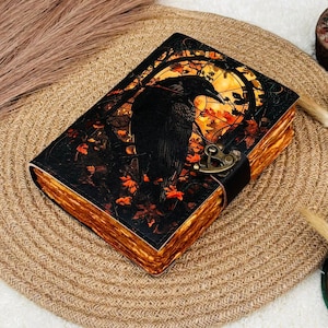 Puede incluir: Un diario encuadernado en cuero con una ilustración de un cuervo en la portada. La portada presenta un cuervo negro sobre un fondo de tonos naranja y amarillo, con un cierre decorativo y una correa de cuero oscuro. Las páginas tienen un aspecto envejecido.