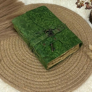 Könnte beinhalten: Ein grünes Notizbuch mit Paisley-Muster, gesichert durch einen Riemen und einen schlüsselförmigen Verschluss. Der Einband des Journals ist tiefgrün, und die Seiten haben ein Vintage-Aussehen. Das Journal liegt auf einem gewebten, kreisförmigen Platzset.