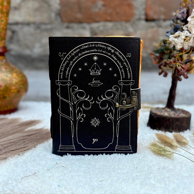 May include: Black leather journal with a decorative design featuring an archway, trees, and stars. The journal has a metal clasp and a decorative border. The pages have a yellowed appearance. The journal is a book.
