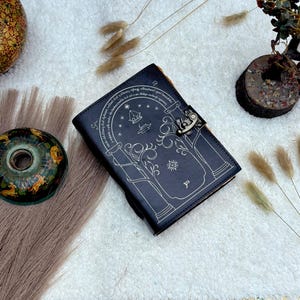 May include: A dark blue leather journal with a decorative silver clasp. The cover features a detailed silver illustration of a tree, stars, and text. The journal is closed and resting on a white surface.