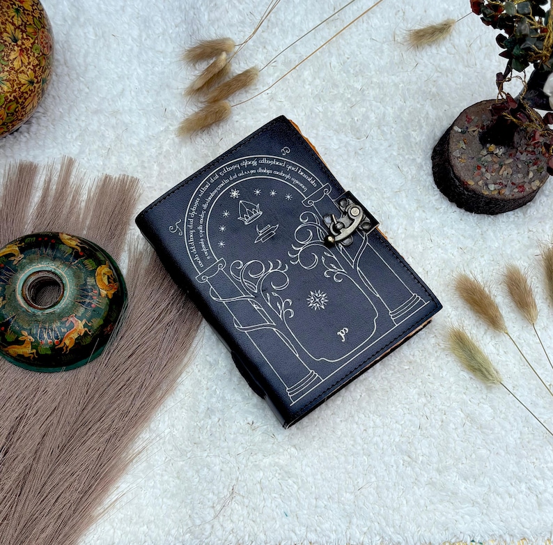 May include: A dark blue leather journal with a decorative silver clasp. The cover features a detailed silver illustration of a tree, stars, and text. The journal is closed and resting on a white surface.