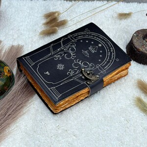 May include: A black leather journal with a decorative tree design and a metal clasp. The pages have a rustic, aged appearance. The cover features a detailed illustration in white, with text around the border.