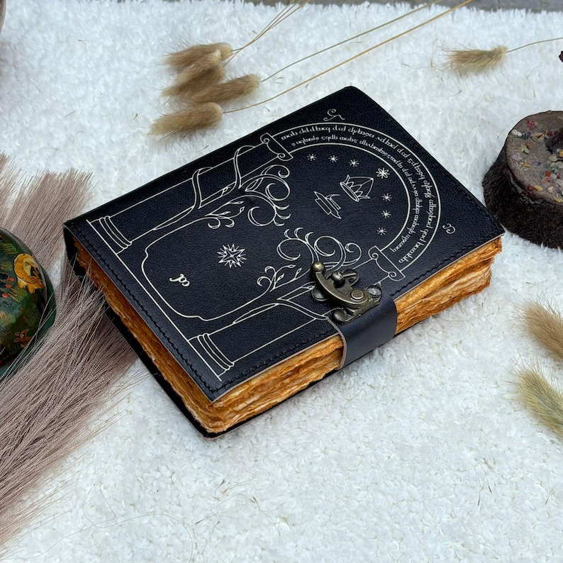 May include: A black leather journal with a decorative tree design and a metal clasp. The pages have a rustic, aged appearance. The cover features a detailed illustration in white, with text around the border.