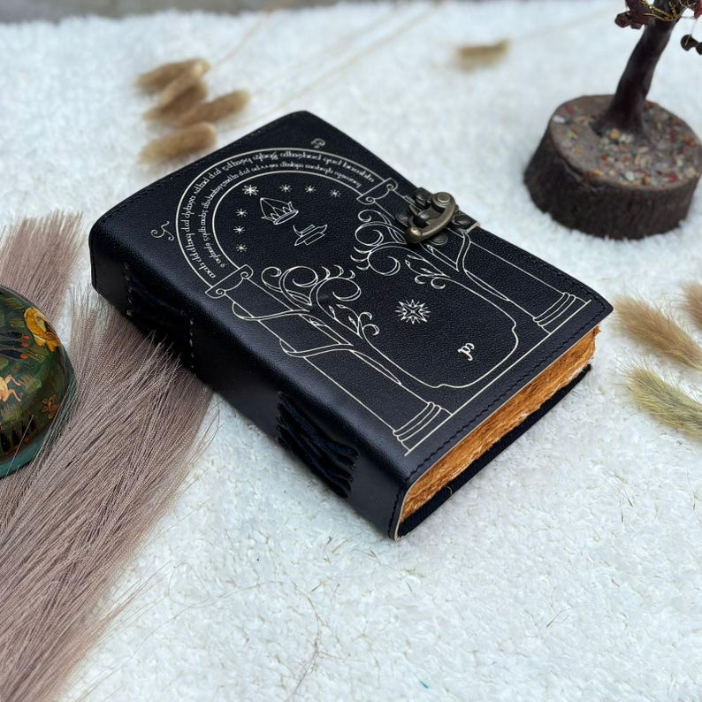 May include: Black leather journal with a decorative design and metal clasp. The cover features a detailed illustration in white, including trees, arches, and text. The pages have a warm, aged appearance.