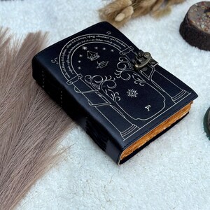 May include: Black leather journal with a decorative white design featuring an arched doorway, stars, and text. The journal has a metal clasp closure and aged-looking pages. A unique, handcrafted notebook.