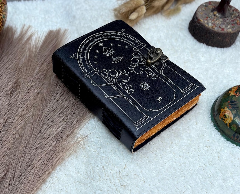May include: Black leather journal with a decorative white design featuring an arched doorway, stars, and text. The journal has a metal clasp closure and aged-looking pages. A unique, handcrafted notebook.