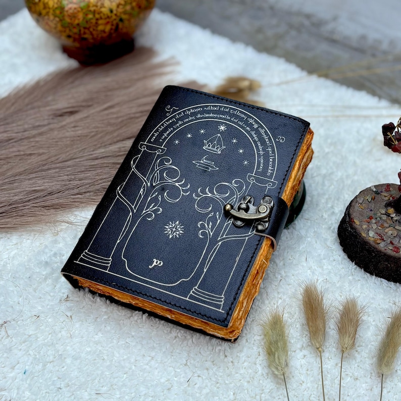 May include: A black leather journal with a decorative design featuring an archway, stars, and a crown. The journal has a metal clasp and aged-looking pages. The cover design is in a light color, contrasting with the dark cover.