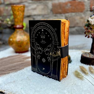 May include: A black leather journal with a silver embossed design featuring an archway, trees, and stars. The journal has a clasp closure and aged, yellowed pages. The edges of the pages are visible, and the journal is bound with a black leather strap.