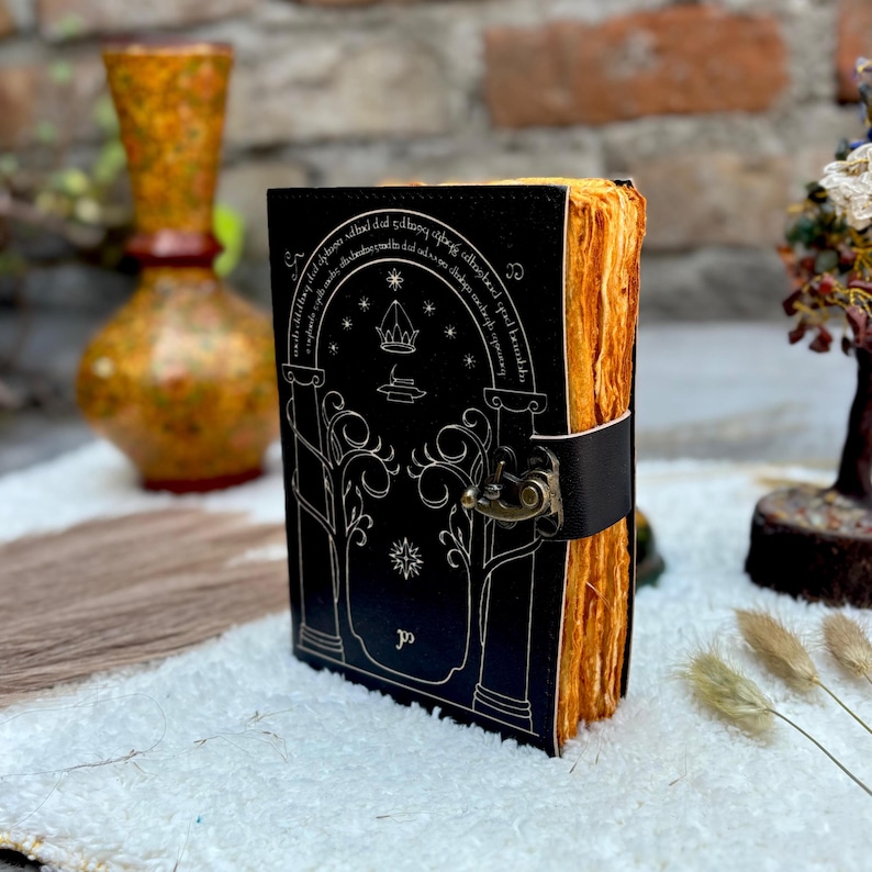 May include: A black leather journal with a silver embossed design featuring an archway, trees, and stars. The journal has a clasp closure and aged, yellowed pages. The edges of the pages are visible, and the journal is bound with a black leather strap.