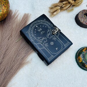 May include: A black leather journal with a decorative design and a metal clasp. The cover features an arched doorway design with stars and text. The journal is closed and lies on a white surface with decorative elements.