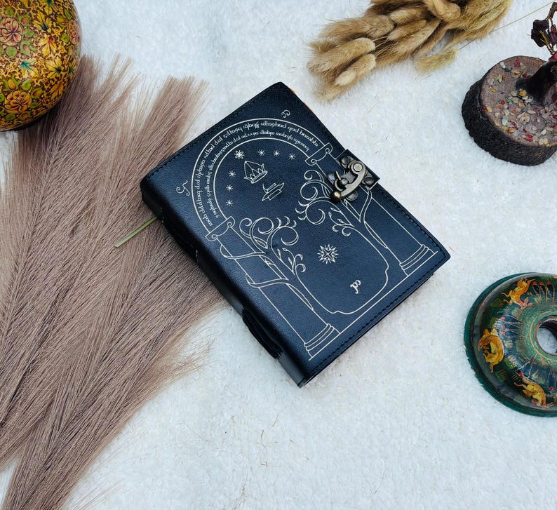 May include: A black leather journal with a decorative design and a metal clasp. The cover features an arched doorway design with stars and text. The journal is closed and lies on a white surface with decorative elements.