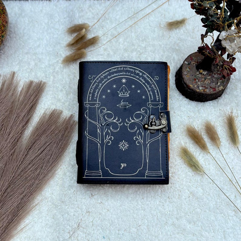 May include: A black leather journal with a silver clasp and a detailed design on the cover. The cover features an arched design with trees, stars, and a crown. The journal is closed and sits on a white surface.