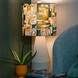 Puede incluir: Una lámpara de mesa con base de cerámica blanca y pantalla rectangular. La pantalla presenta un patrón geométrico de caras en tonos amarillos, verdes y azules. Una planta de aloe vera en una maceta naranja está cerca.