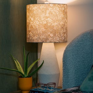 Peut inclure: Une lampe de chevet avec un abat-jour à motifs floraux est posée sur une table de nuit en bois. La lampe a un pied blanc et se trouve à côté d'une plante d'aloe vera en pot orange. Une paire de lunettes repose sur un livre.