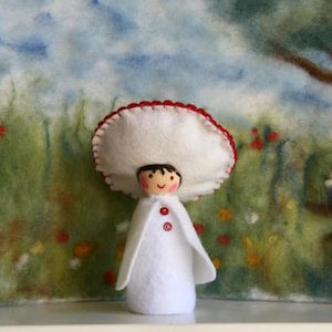 Könnte beinhalten: Eine skurrile Filzpuppe mit einem Pilzhut, roter Bordüre und einem weißen Umhang. Die Puppe hat ein bemaltes Gesicht und rote Knöpfe. Der Hintergrund zeigt eine gemalte Landschaft mit einem Baum und einem Zwerg.