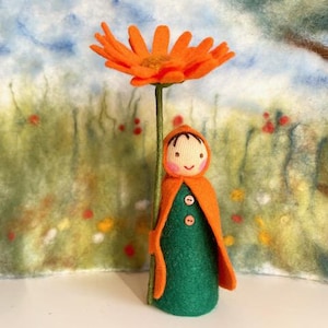 Könnte beinhalten: Eine Filzpuppe mit grünem Kleid und orangefarbenem Umhang hält eine große orangefarbene Filzblume.