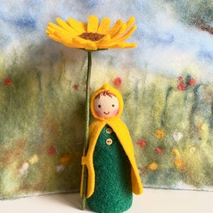 Könnte beinhalten: Eine Filzpuppe mit einem gelben Kapuzenmantel und einem grünen Kleid, die eine gelbe Filzsonnenblume hält.