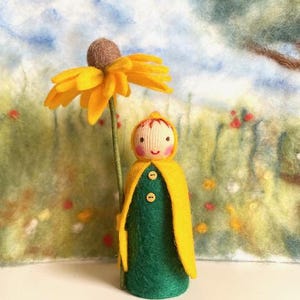 Könnte beinhalten: Eine Filzpuppe, die einen gelben Umhang mit Kapuze und ein grünes Kleid trägt, hält eine gelbe Filzblume mit braunem Zentrum. Die Puppe steht vor einem Filzhintergrund mit einer grünen, gelben und blauen Landschaft.