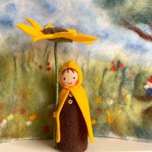 Könnte beinhalten: Eine Filzpuppe mit einem gelben Kapuzenumhang hält eine gelbe Filzsonnenblume. Die Puppe hat braunes Haar und ein lächelndes Gesicht. Der Hintergrund ist eine Filzlandschaft mit einem Baum und einem kleinen Kobold.