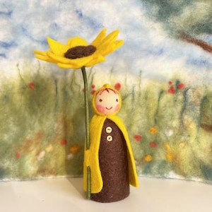 Könnte beinhalten: Eine Filzpuppe mit einem gelben Kapuzenumhang hält eine gelbe Filzsonnenblume. Die Puppe hat einen braunen Filzkörper und ein lächelndes Gesicht mit rosa Wangen. Die Puppe steht vor einem Filzhintergrund mit einer grün-gelb-blauen Landschaft.