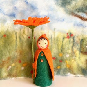 Könnte beinhalten: Handgefertigte Filzpuppe mit grünem Kleid und orangefarbenem Kapuzenmantel, die eine große orangefarbene Blume hält. Die Puppe hat ein lächelndes Gesicht. Der Hintergrund ist eine Filzlandschaft mit einem Baum und Blumen.
