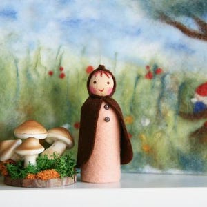 Könnte beinhalten: Eine Filzpuppe mit braunem Umhang und lächelndem Gesicht steht neben einer Gruppe von Pilzfiguren. Der Hintergrund zeigt eine Filzlandschaft mit einem Baum und einem Zwerg. Die Puppe ist etwa 13 cm groß.
