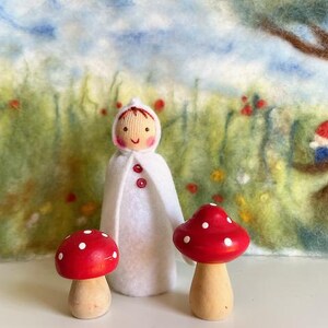Könnte beinhalten: Eine weiße Filzpuppe mit einem roten Knopf und einem roten lächelnden Gesicht steht vor einer Filzkulisse mit einem Baum und Blumen. Zwei rote und weiße Pilzfiguren stehen vor der Puppe.