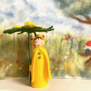 Könnte beinhalten: Eine Filzpuppe, die einen gelben Regenmantel trägt und einen grünen Filzschirm hält. Die Puppe hat ein lächelndes Gesicht mit braunen Augen und einem roten Mund. Die Puppe steht vor einem Filzhintergrund mit einem Baum und Blumen.