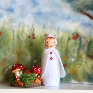 Könnte beinhalten: Eine Filzpuppe in einem weißen Umhang mit roten Knöpfen und passender Mütze steht vor einer skurrilen Kulisse. Eine Gruppe von rotkappigen Pilzen sitzt auf einem Holzsockel. Der Hintergrund zeigt eine gemalte Wiesenlandschaft.