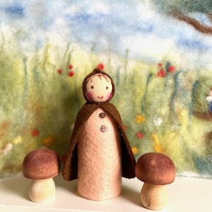 Könnte beinhalten: Eine Filzpuppe mit einem braunen Umhang und einem rosa Kleid steht zwischen zwei braunen Pilzfiguren vor einem grün-blauen Filzhintergrund.