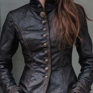 Puede incluir: Chaqueta de cuero negro con cuello alto y botones dorados decorativos. La chaqueta presenta costuras marrones a lo largo de las costuras y puños. Una prenda elegante y entallada.