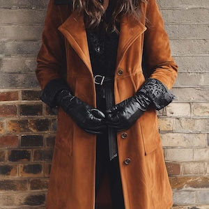 May include: A long, brown suede coat with a black collar and button closure. The coat is paired with black leather gloves with lace cuffs and a black belt. The outfit is set against a brick wall.