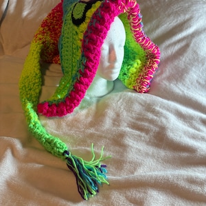 May include: A brightly coloured, handmade crochet hat with long, ear-like extensions. The hat features vibrant neon green, pink, yellow, and blue yarn. A tassel of blue, green, and purple yarn hangs from the end. The hat is displayed on a white mannequin head.