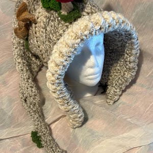 May include: A whimsical, handmade crochet hat in shades of grey and beige, adorned with red and white mushroom accents and green leaves. The hat features a long, trailing stem with a green tassel, creating a unique and playful design. The hat is designed to fit a head.