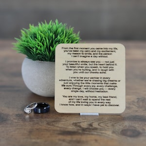 May include: A wedding ring set with a diamond engagement ring and a black band ring, displayed with a small potted plant and a wooden plaque with a vow. The plaque has a poem about love and commitment.