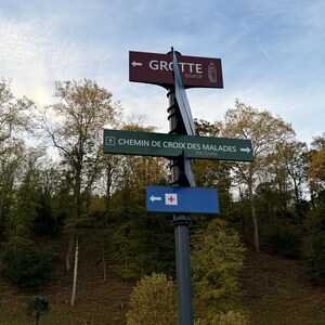 Puede incluir: Un poste de se&ntilde;alizaci&oacute;n met&aacute;lico con tres se&ntilde;ales direccionales. La se&ntilde;al superior es granate con texto blanco "GROTTE" y una ilustraci&oacute;n. La se&ntilde;al del medio es verde con texto blanco "CHEMIN DE CROIX DES MALADES". La se&ntilde;al inferior es azul con una cruz blanca.