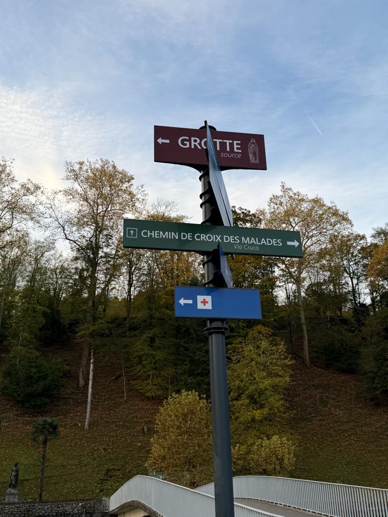 Puede incluir: Un poste de se&ntilde;alizaci&oacute;n met&aacute;lico con tres se&ntilde;ales direccionales. La se&ntilde;al superior es granate con texto blanco "GROTTE" y una ilustraci&oacute;n. La se&ntilde;al del medio es verde con texto blanco "CHEMIN DE CROIX DES MALADES". La se&ntilde;al inferior es azul con una cruz blanca.
