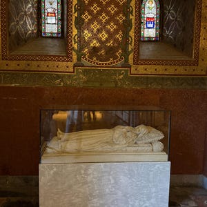 Puede incluir: Una escultura de m&aacute;rmol de una persona acostada, encerrada en una caja de cristal, sobre un pedestal gris claro. El fondo presenta vidrieras y paredes de mosaico dorado y rojo ornamentadas.
