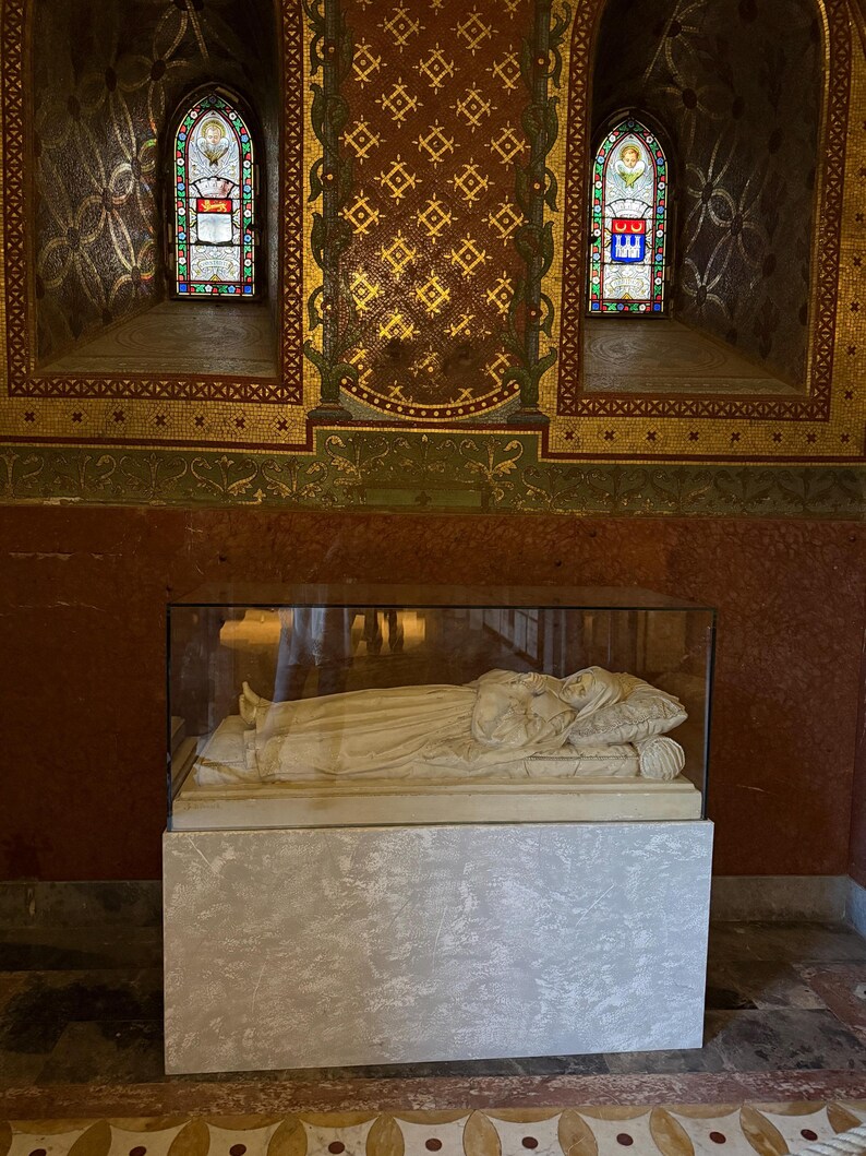 Puede incluir: Una escultura de m&aacute;rmol de una persona acostada, encerrada en una caja de cristal, sobre un pedestal gris claro. El fondo presenta vidrieras y paredes de mosaico dorado y rojo ornamentadas.